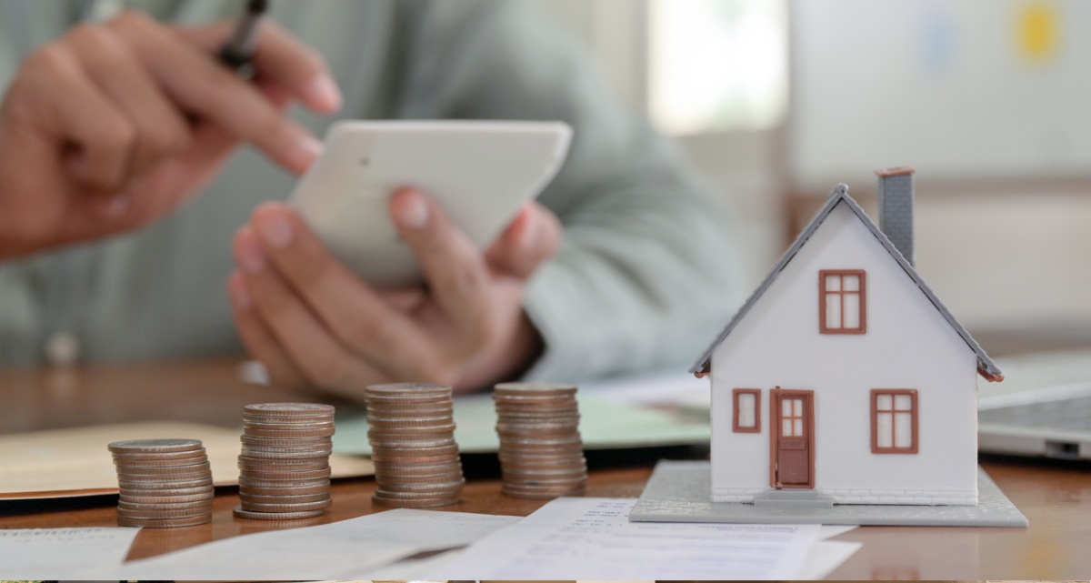 A model house and a stack of coins on a table with a pair of hands holding a calculator in the background