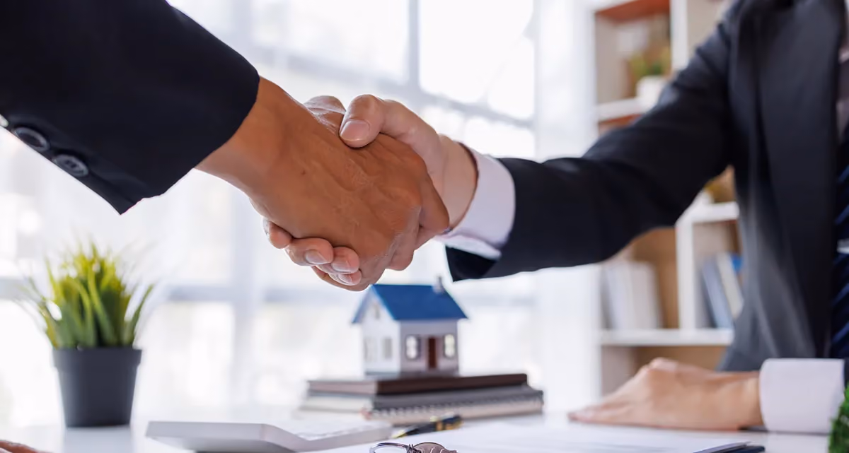 2 men shaking hands with a calculator and a miniature model house on a stack of books in the background