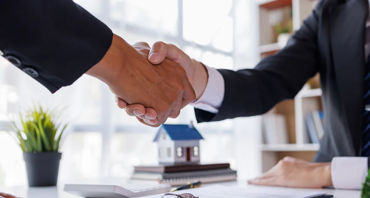 2 men shaking hands with a calculator and a miniature model house on a stack of books in the background