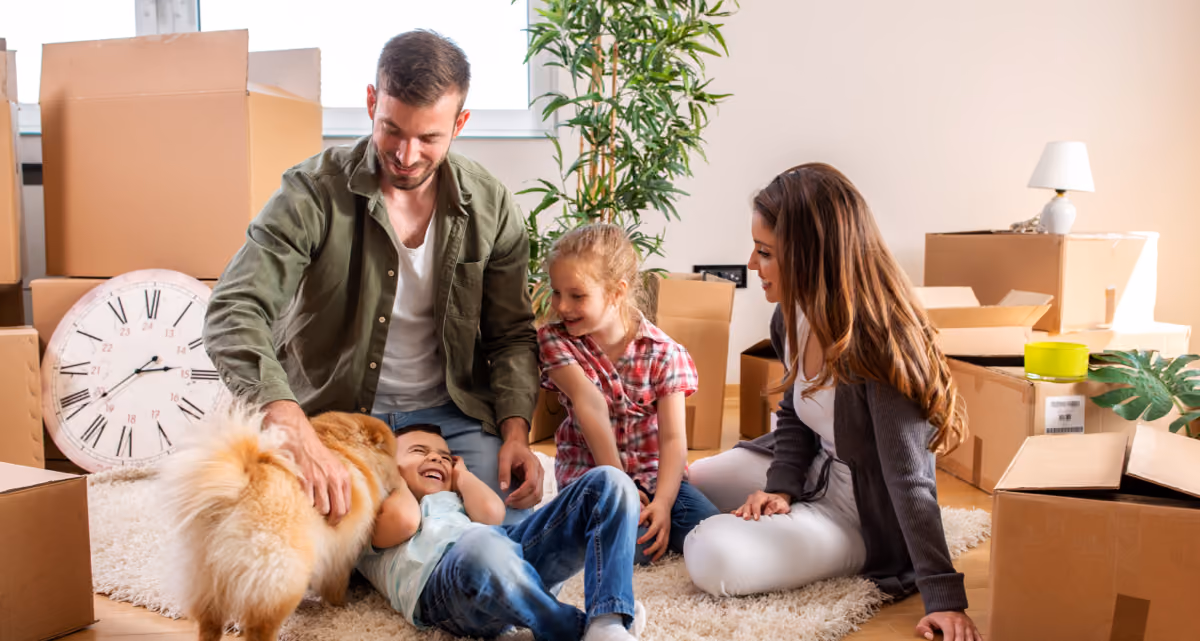 A family with a dog sitting on the floor surrounded by moving boxes