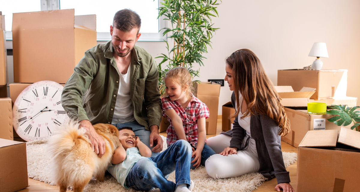 A family with a dog sitting on the floor surrounded by moving boxes