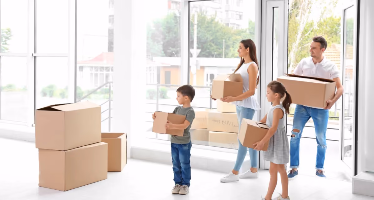 Two adults and two children carrying cardboard moving boxes into a building