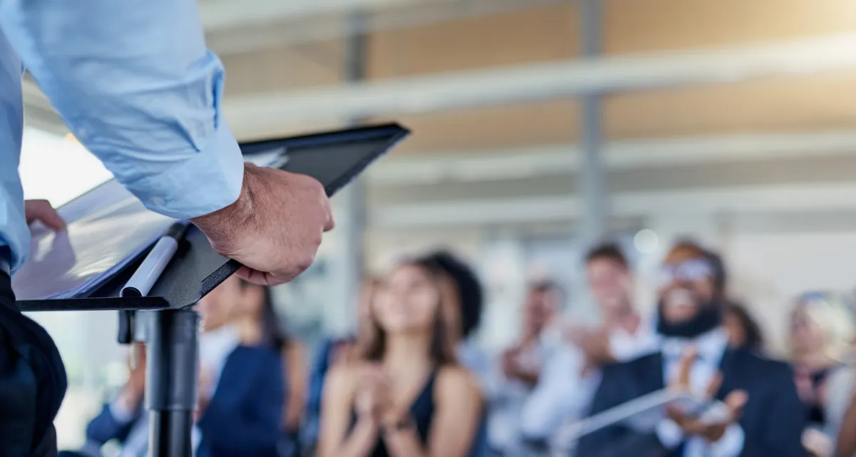 An audience looking on as a person delivers a speech from a podium