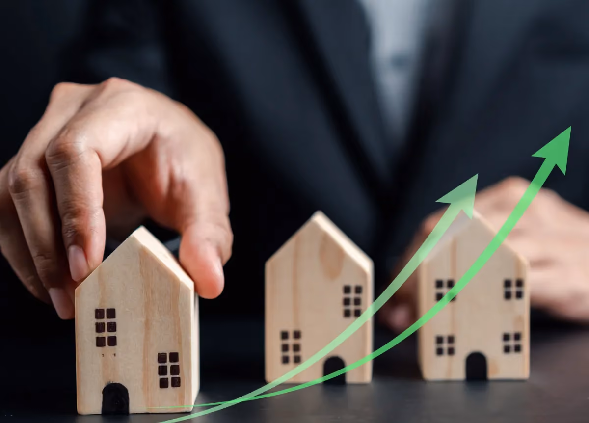 A person picking up a model house with some rising green arrows over the top