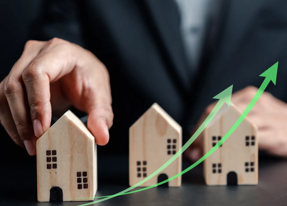 A person picking up a model house with some rising green arrows over the top
