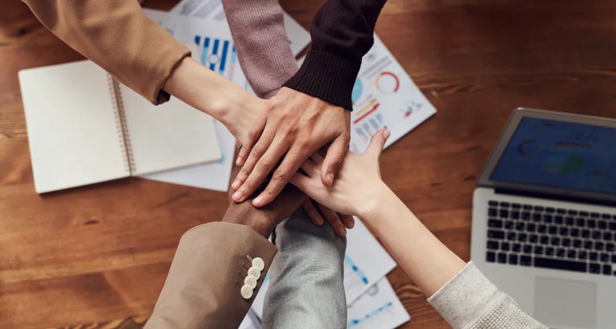 A group of people stacking their hands over a desk with documents and a laptop