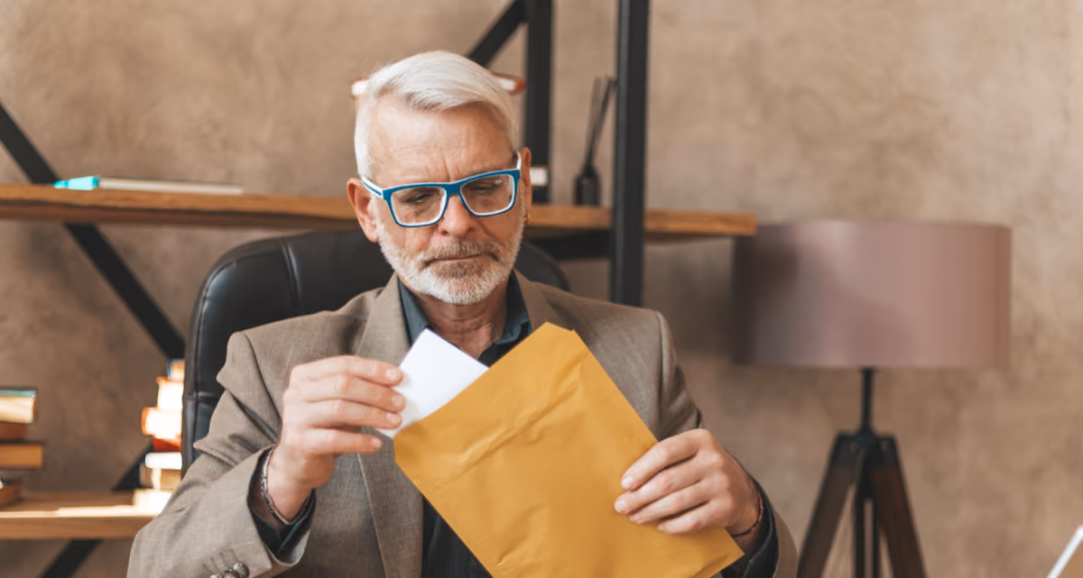 A seated man in a suit and glasses taking a large letter out of an envelope
