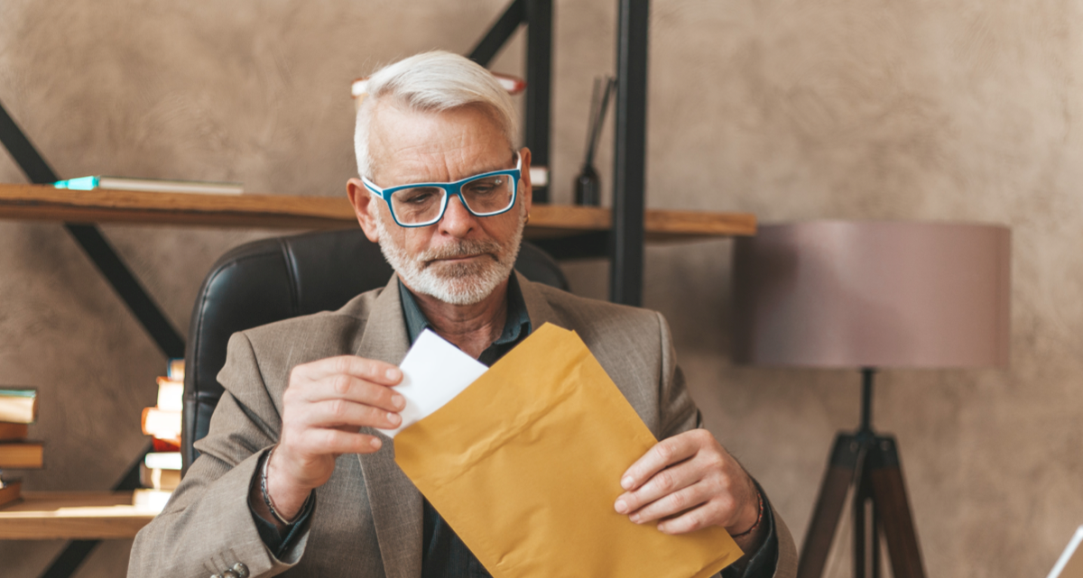 A seated man in a suit and glasses taking a large letter out of an envelope