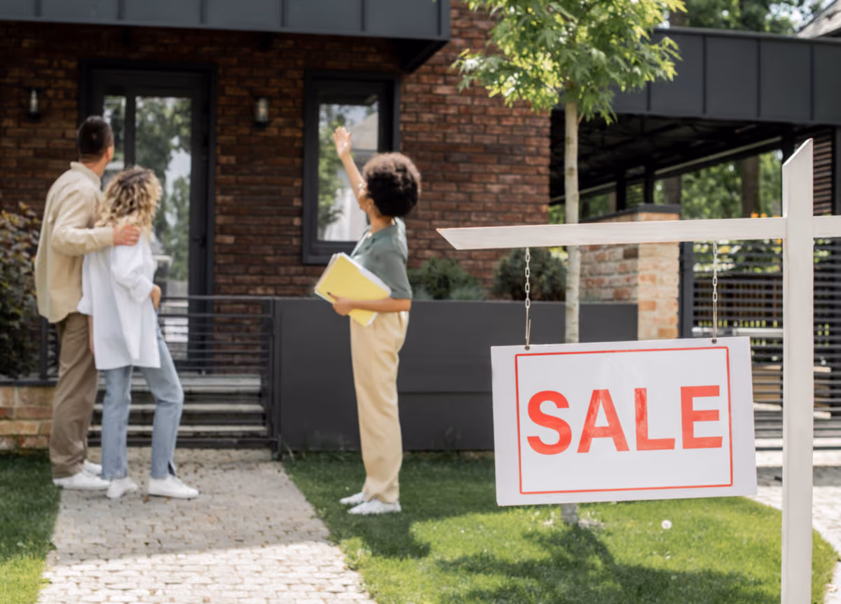 Real estate agent showing house to hugging couple near “for sale” signboard