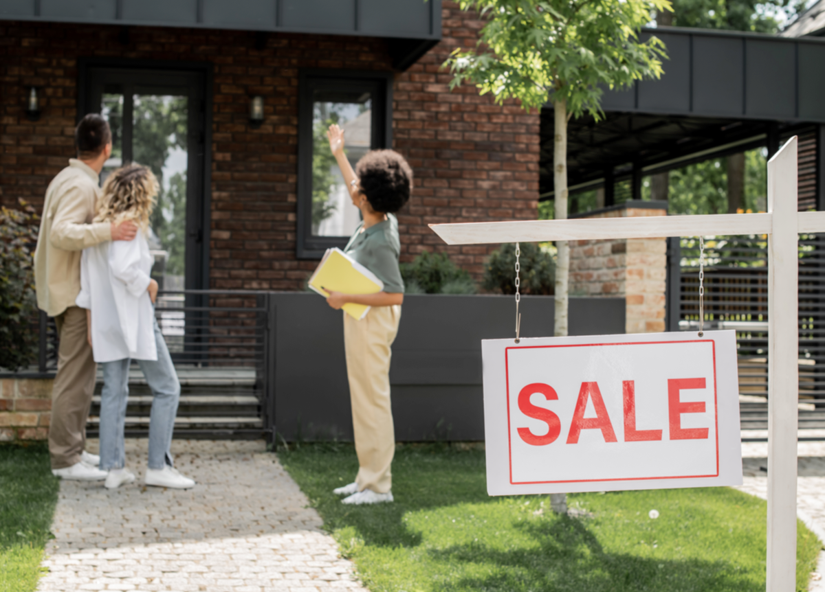Real estate agent showing house to hugging couple near “for sale” signboard