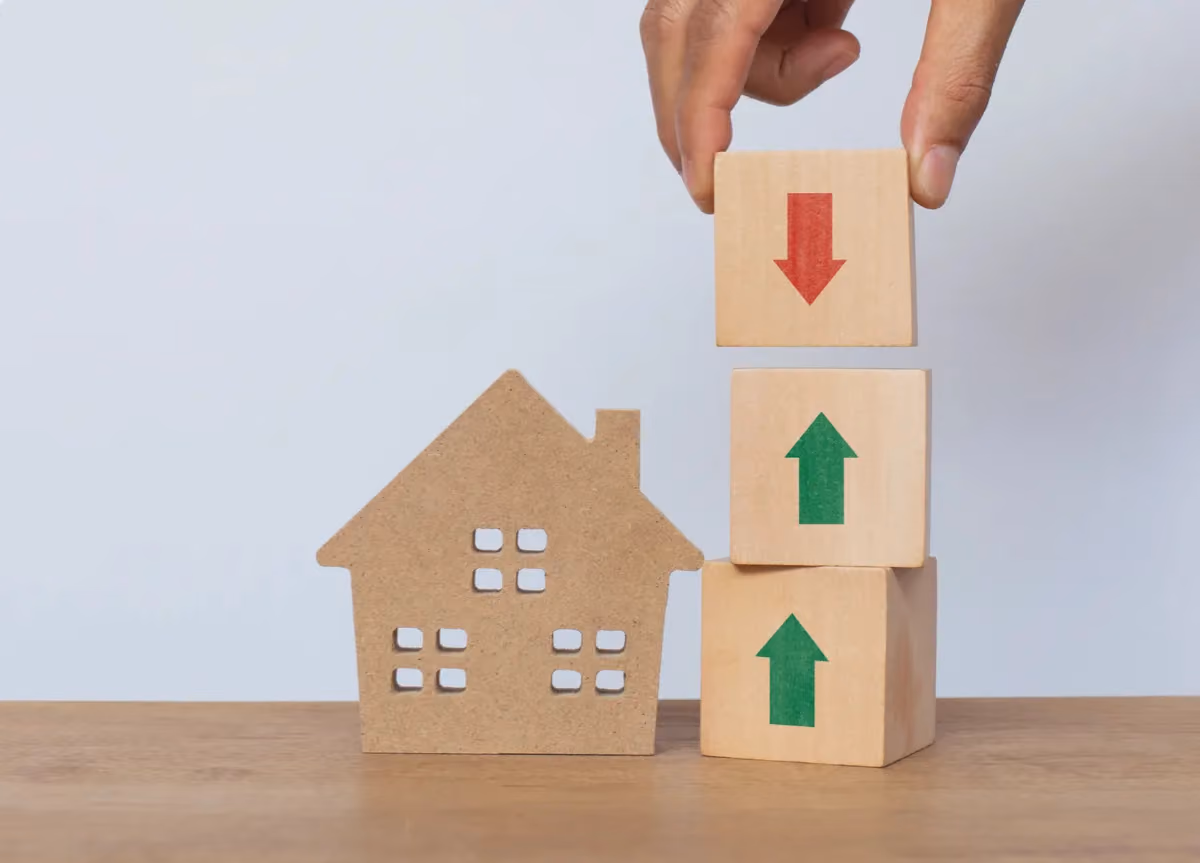 Hand arranging wooden cube stack with one arrow pointing up and two arrows down, next to a wooden house model on white background