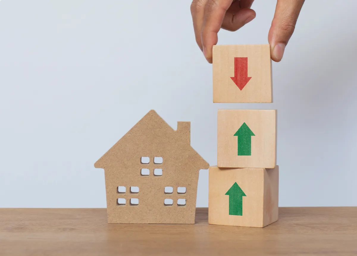 Hand arranging wooden cube stack with one arrow pointing up and two arrows down, next to a wooden house model on white background