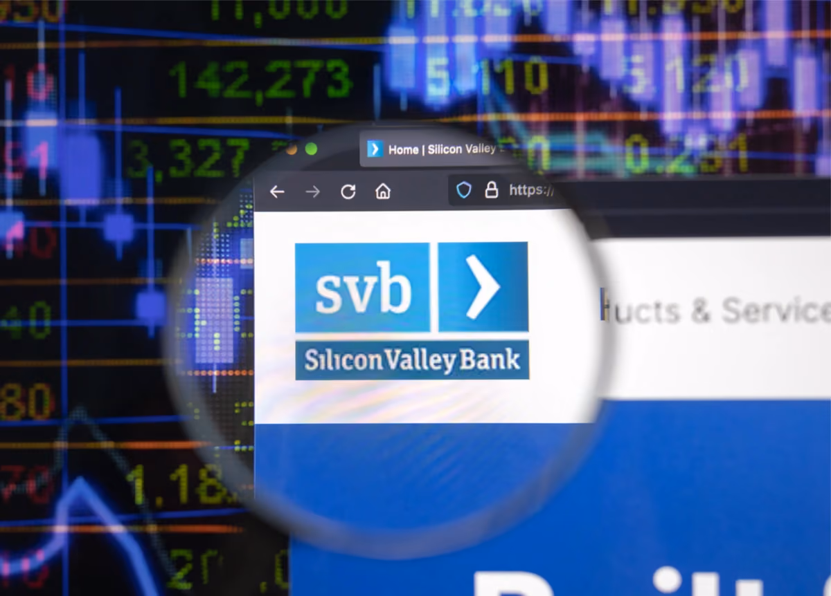 Silicon Valley Bank company logo seen on a computer screen through a magnifying glass with blurry stock market developments in the background.