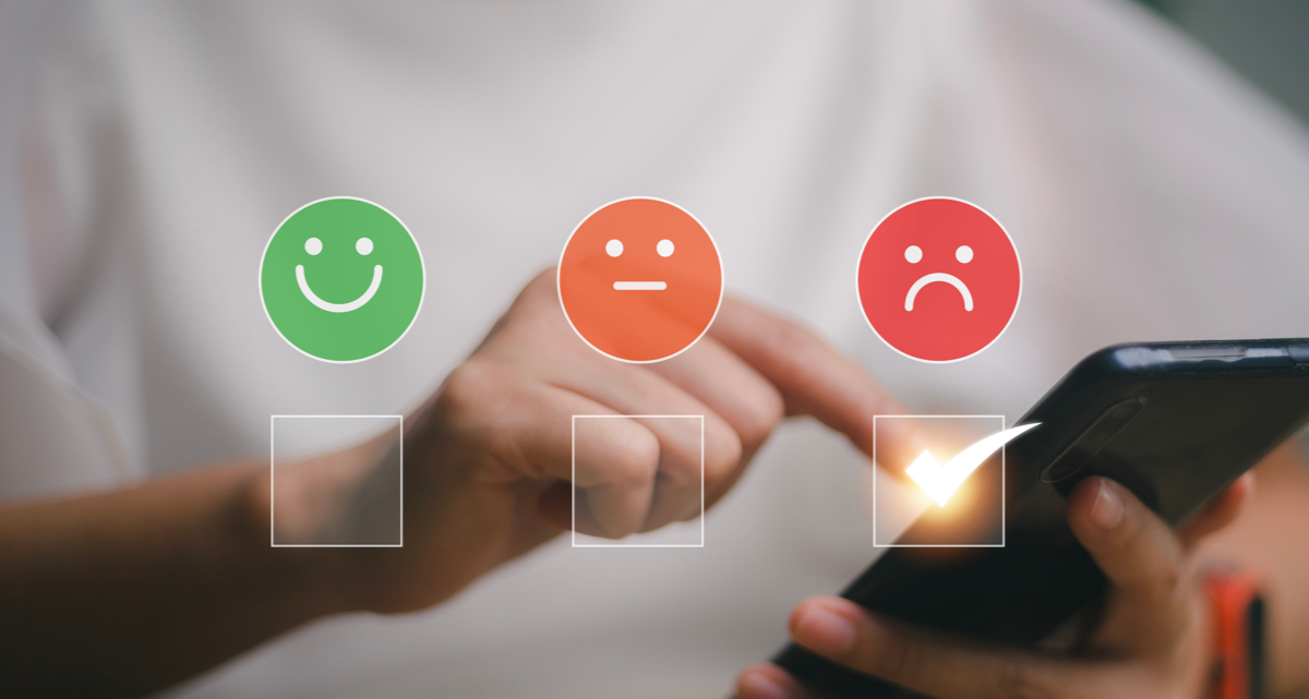 A closeup of a person tapping their smartphone screen with a green smiley face, an orange neutral face and a red frowning face over it
