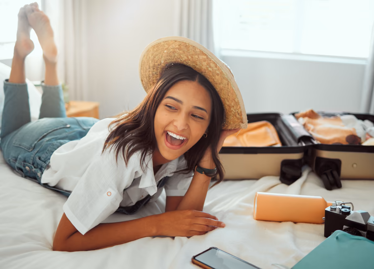 Smiling woman relaxing on hotel bed with suitcase, ready for summer vacation