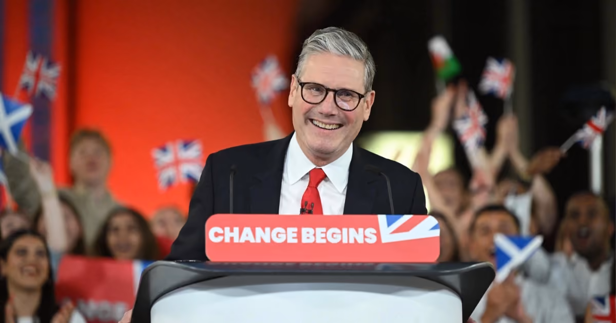 Sir Kier Starmer standing on a podium and smiling with supporters waving flags in the background. The front of the podium reads "change begins".