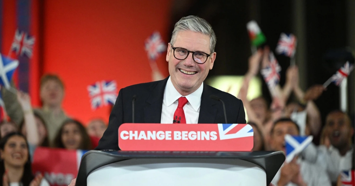 Sir Kier Starmer standing on a podium and smiling with supporters waving flags in the background. The front of the podium reads "change begins".