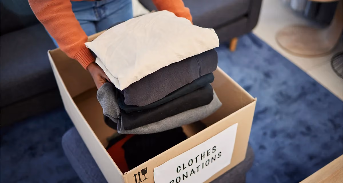 A person putting a stack of winter clothes into a box marked "Clothes donations"