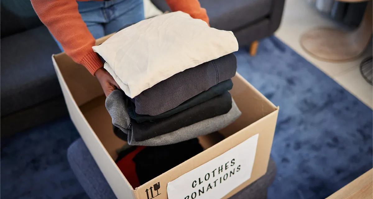 A person putting a stack of winter clothes into a box marked "Clothes donations"