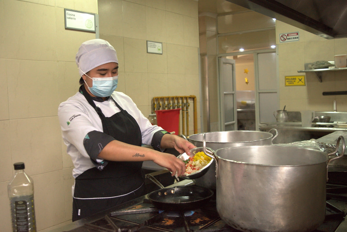 Cocinera prepara alimentos junto a ollas industriales en una cocina institucional
