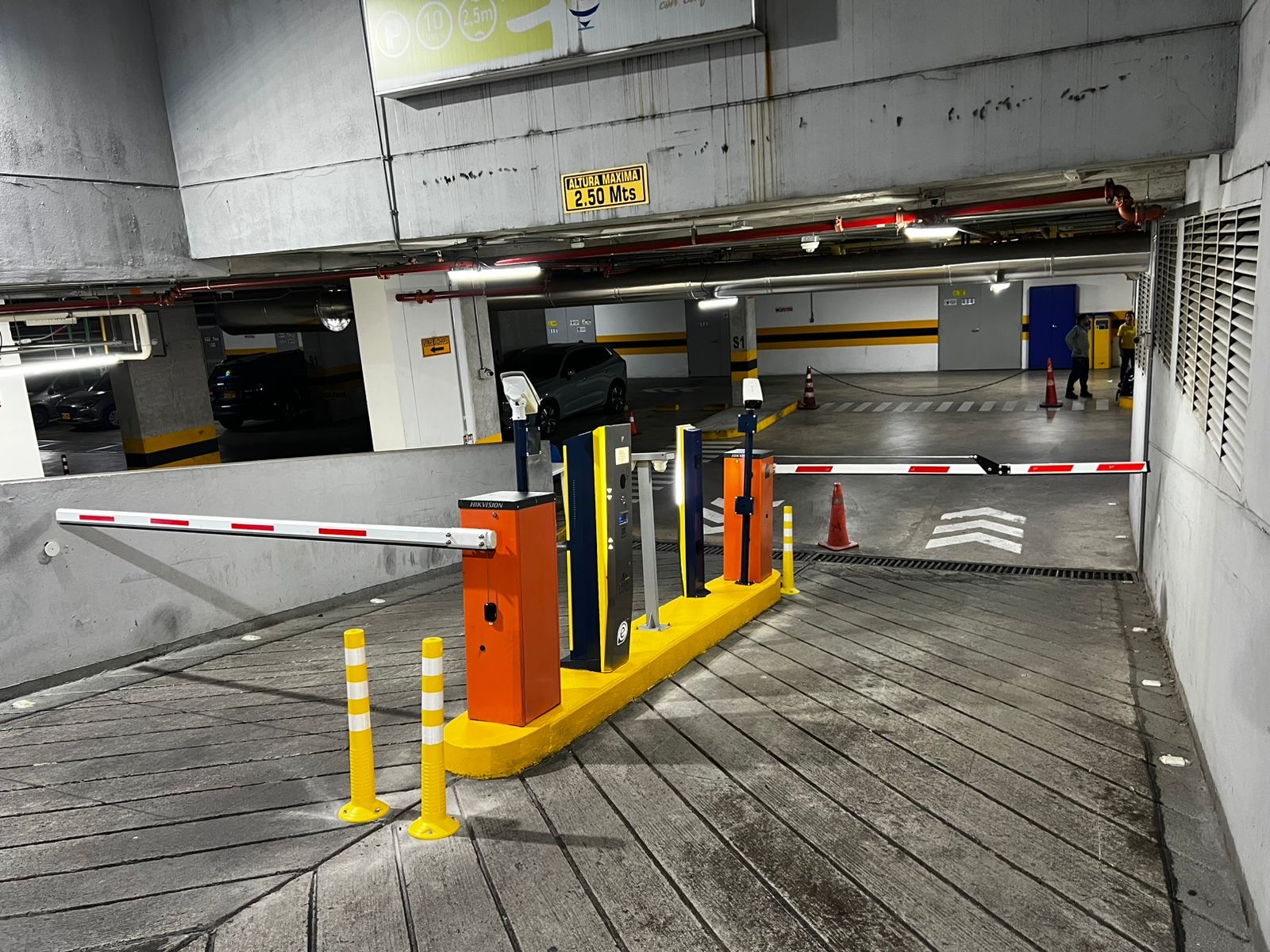 Acceso a un parqueadero cubierto con barreras, equipos de entrada y salida y rampa señalizada.