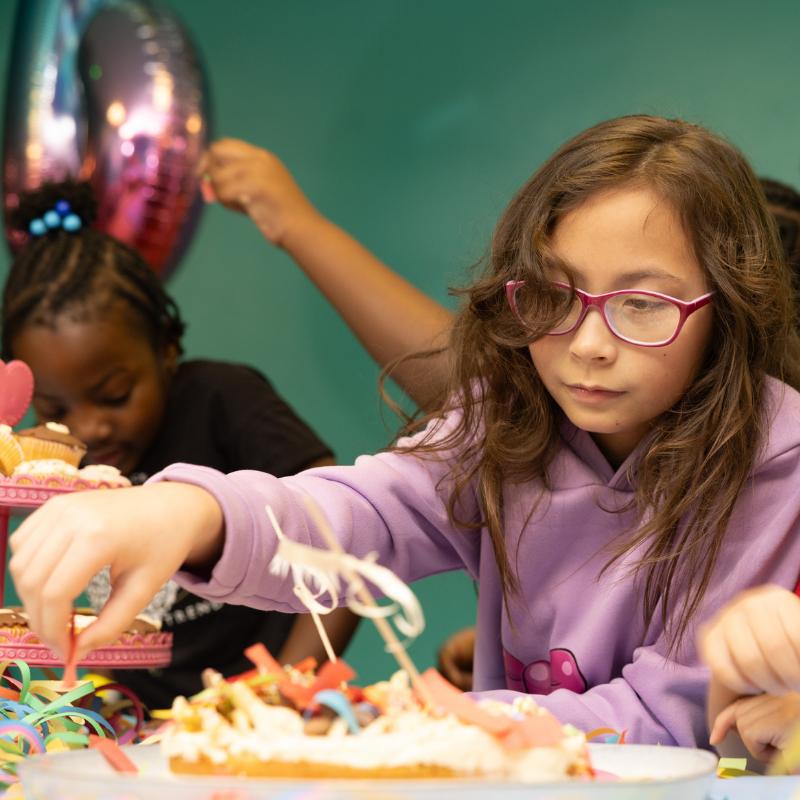 Girl in purple hoodie decorating cake with colorful streamers at party