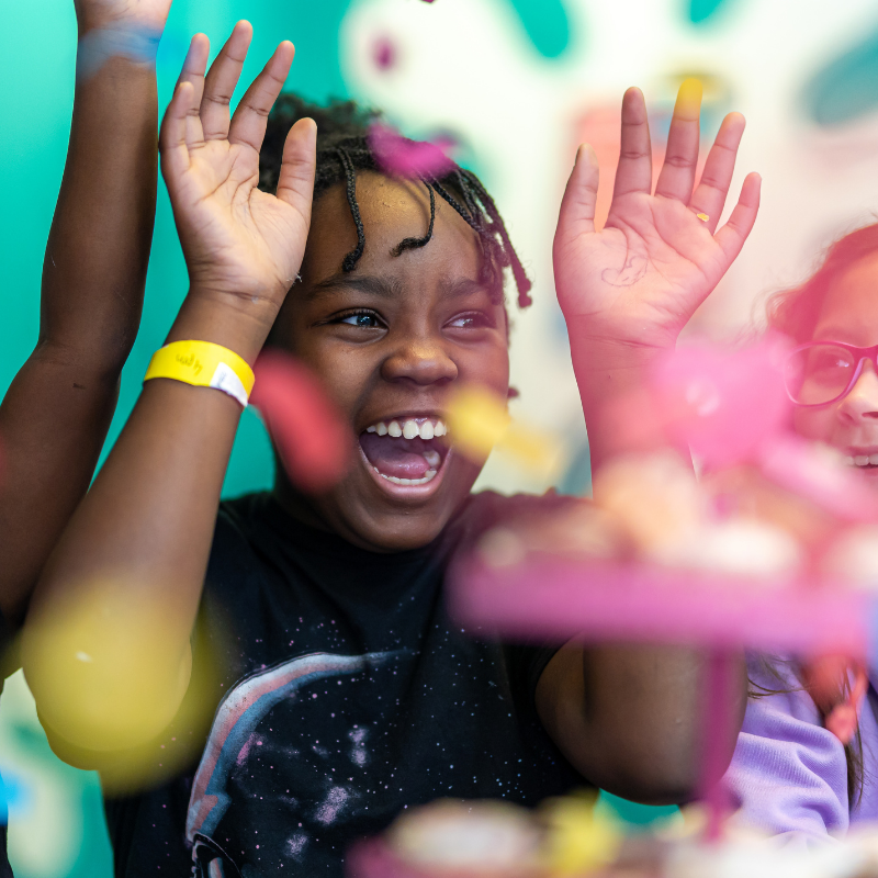 Excited child raising hands, laughing amid colorful blurred confetti
