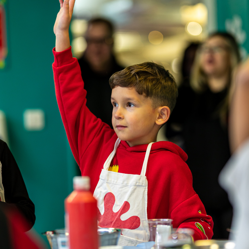 Young boy in red hoodie raising hand during cooking class