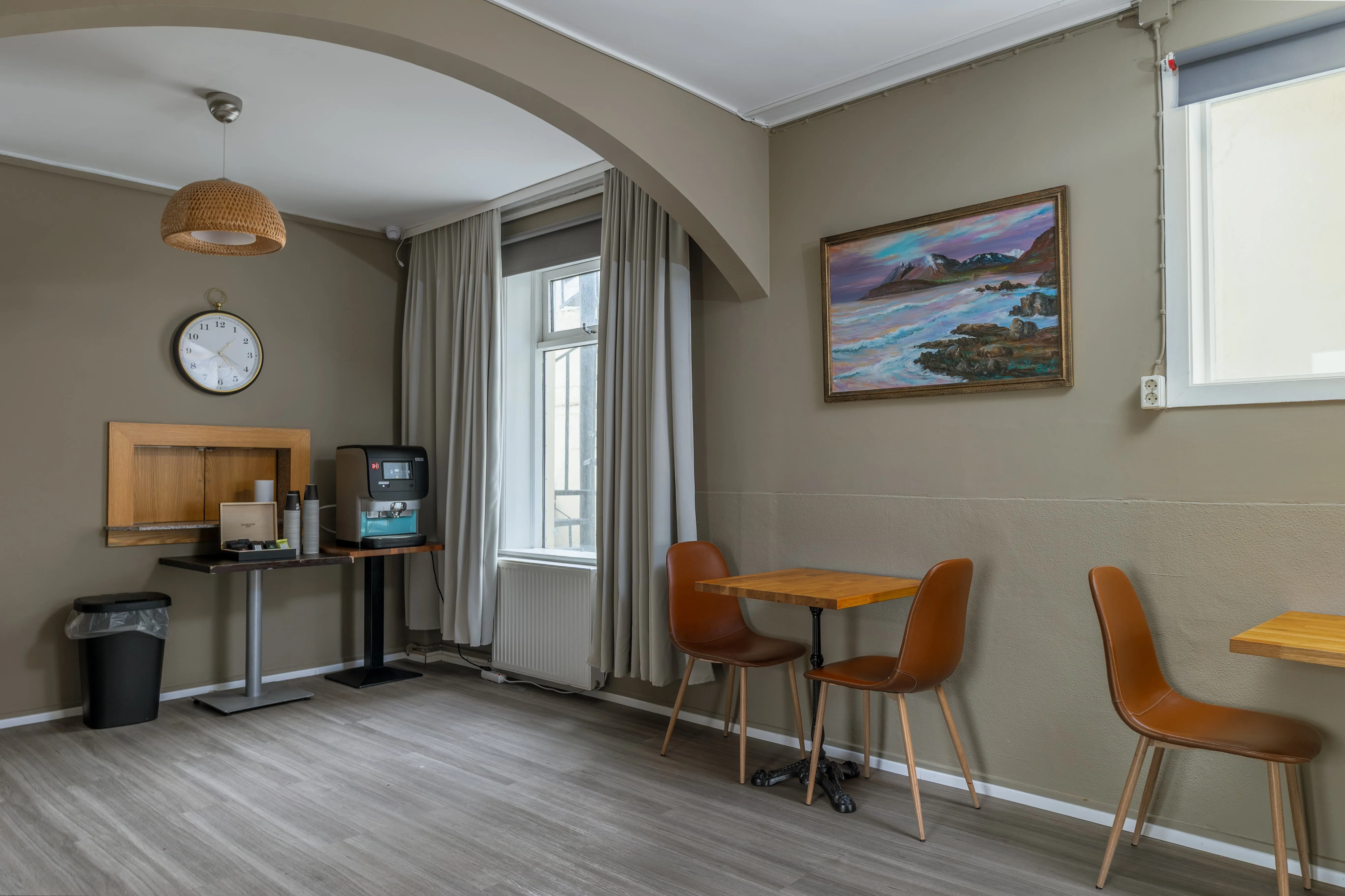 Common area at Old Town Reykjavik with two small wooden tables, three brown chairs, a coffee machine with complimentary tea & coffee for guests, a wall clock, and a framed painting of a seascape with mountains.