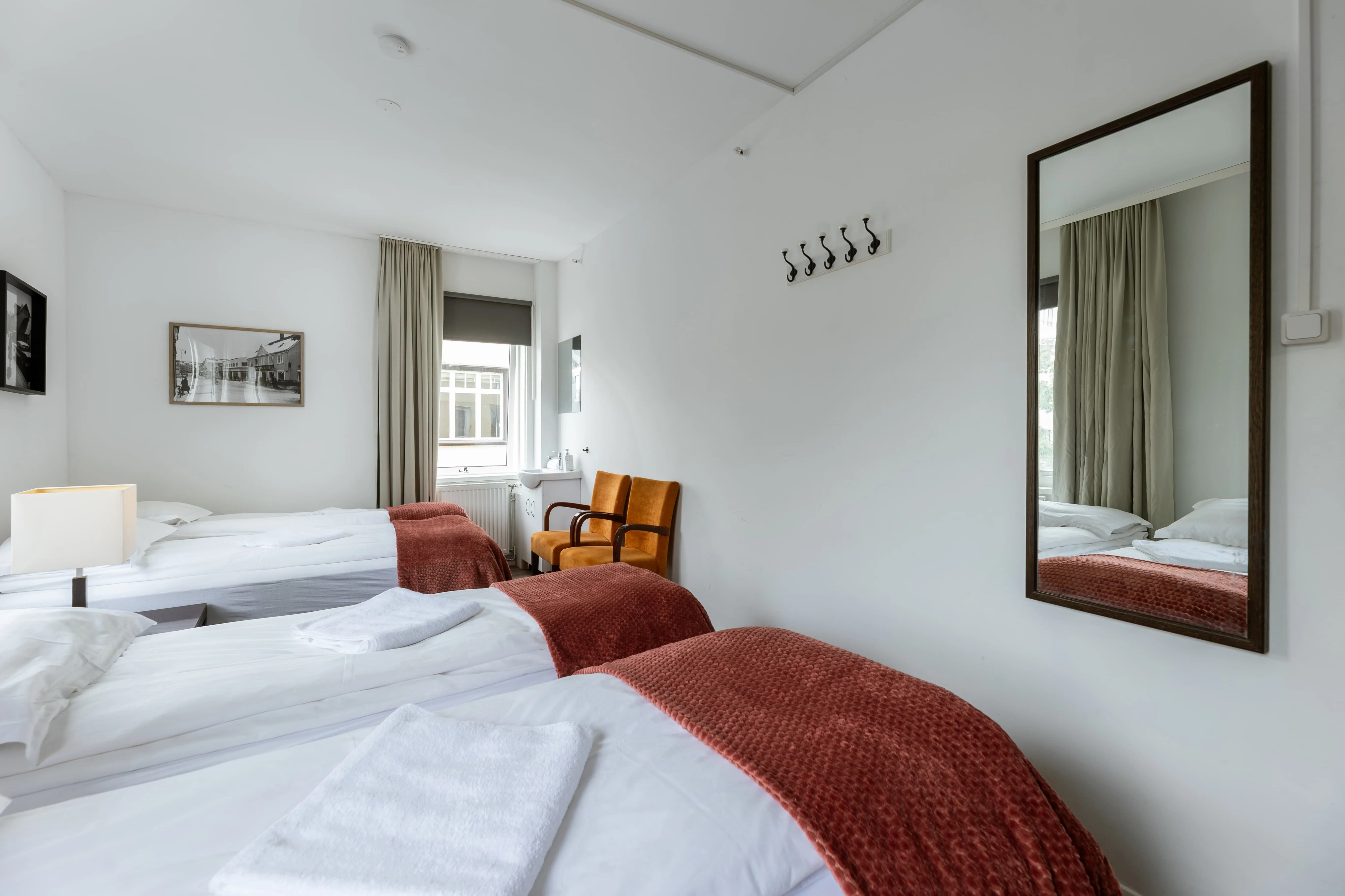 Bright minimalist hotel room at Old Town Reykjavik with three single beds, white bedding, rust-colored throws, two orange chairs, and a large wall mirror.