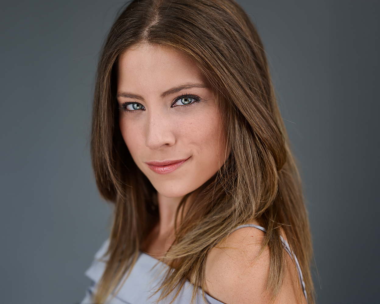 Professional headshot of Heather, facing camera with slight smile, long brown hair over shoulder, gray background.