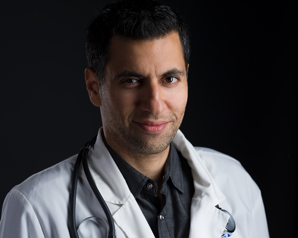 Professional headshot of a confident man wearing a white doctor’s coat and stethoscope, looking directly at the camera with a slight smile against a dark background.