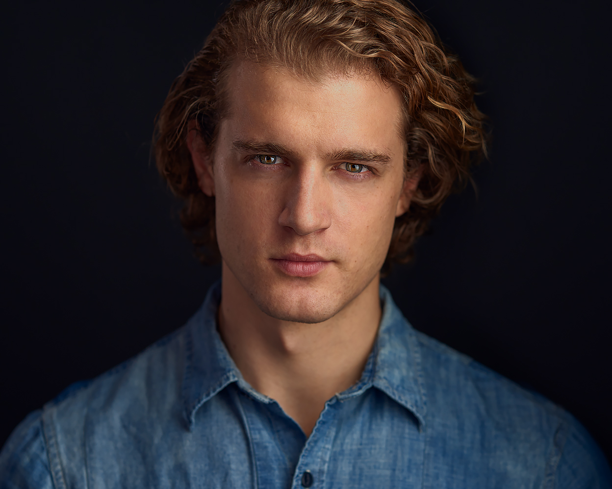 Confident man in a denim shirt against a dark background, making direct eye contact with a neutral expression.