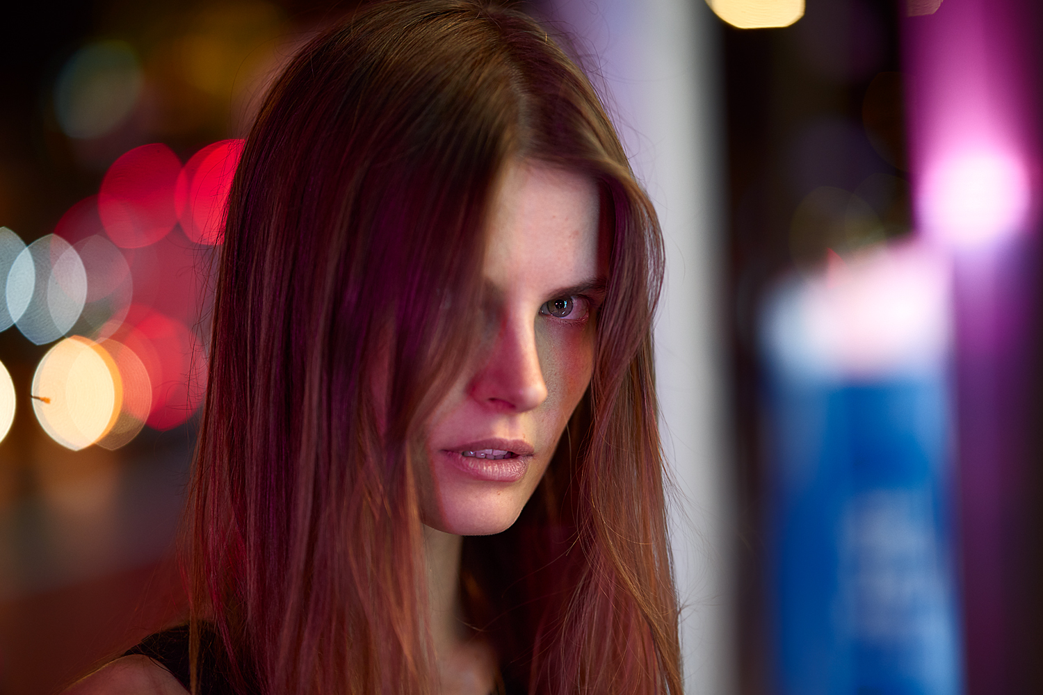 Portrait of a woman with long, straight red-toned hair under vibrant magenta and violet lighting. She glances off-camera with an intense expression, surrounded by neon city glow.