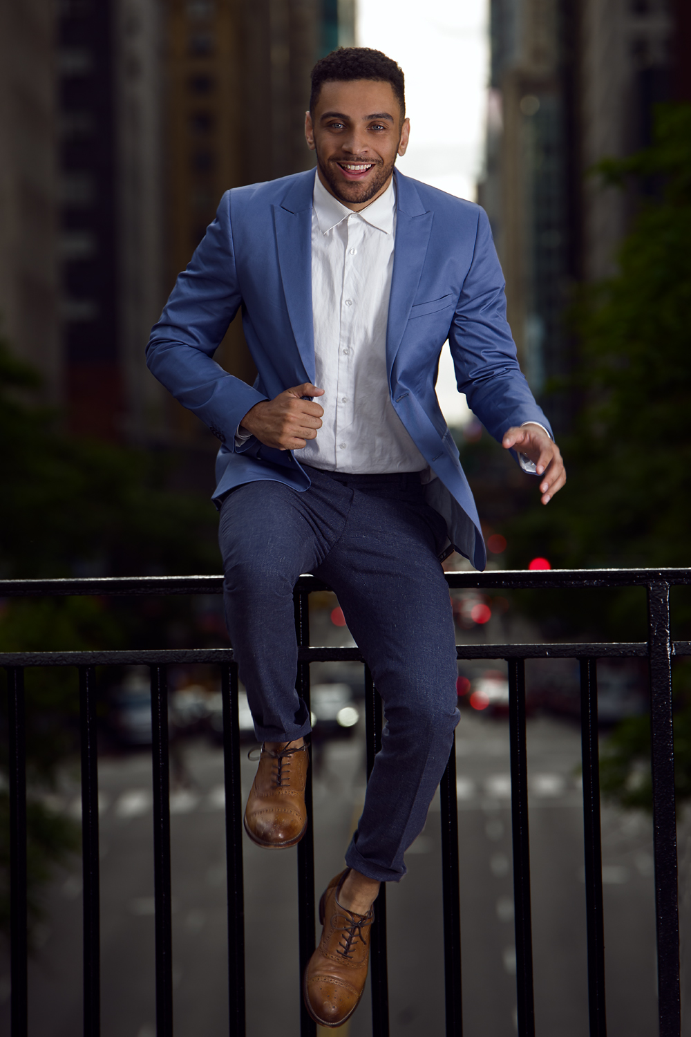Portrait of a man in a blue blazer and brown dress shoes, captured mid-jump above a city street. He smiles confidently, suspended in motion, with blurred buildings in the background.