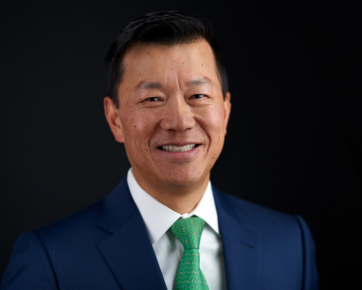 Professional headshot of a man in a dark blue suit, white shirt, and green tie, smiling confidently against a dark background.