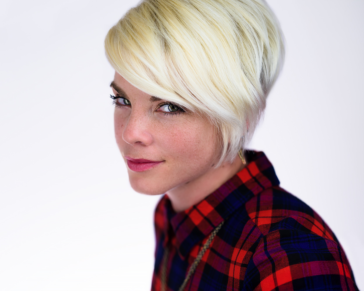 Blonde woman in a red plaid shirt with a short angled haircut, gazing confidently at the camera against a clean white background.