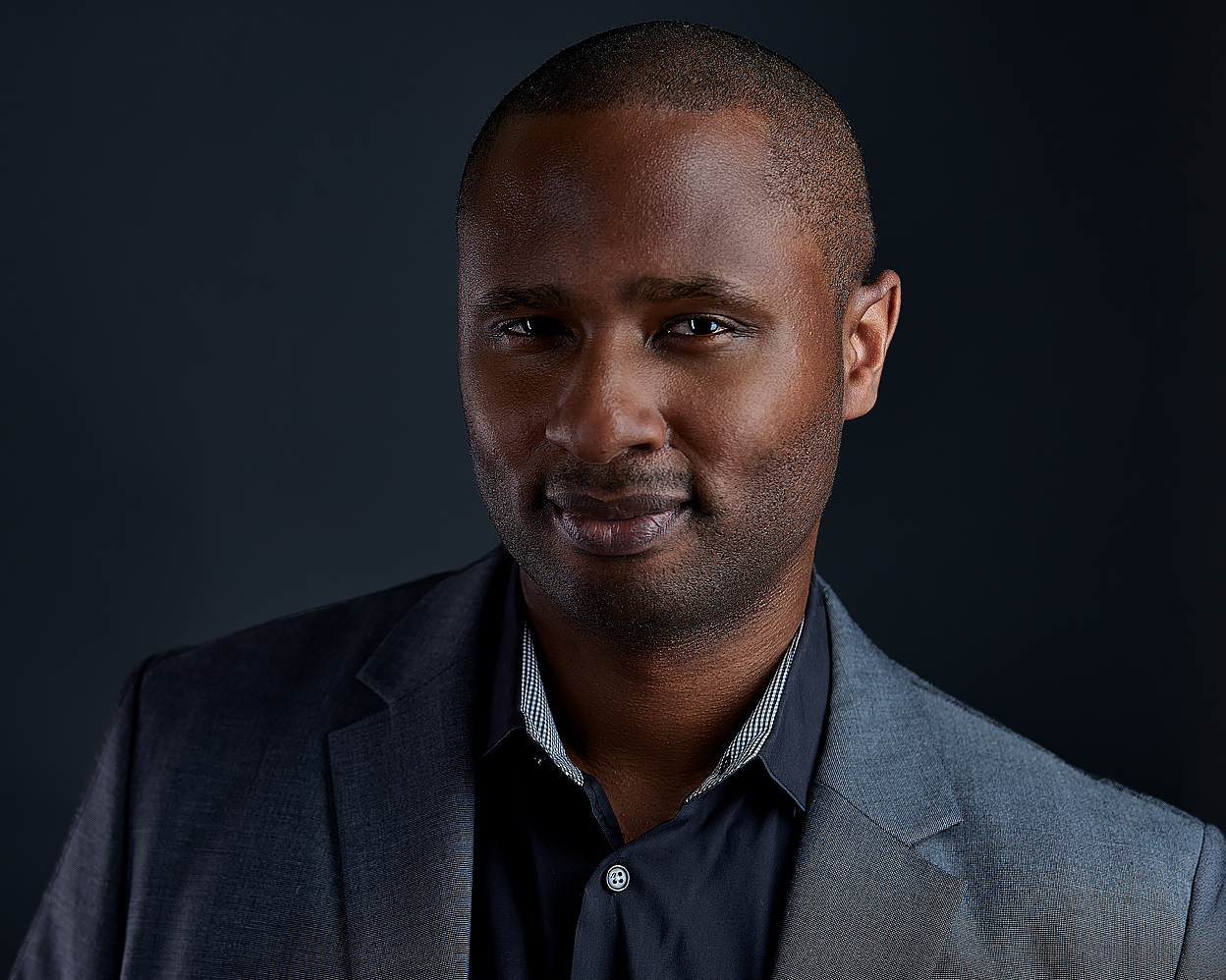 Man with closely cropped hair wearing a gray blazer and black dress shirt, looking confidently at the camera against a dark studio background.