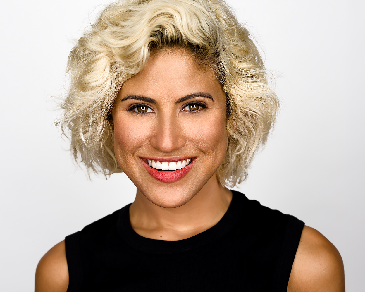 Woman with short platinum blonde curly hair wearing a black sleeveless top, smiling warmly in front of a bright white background.