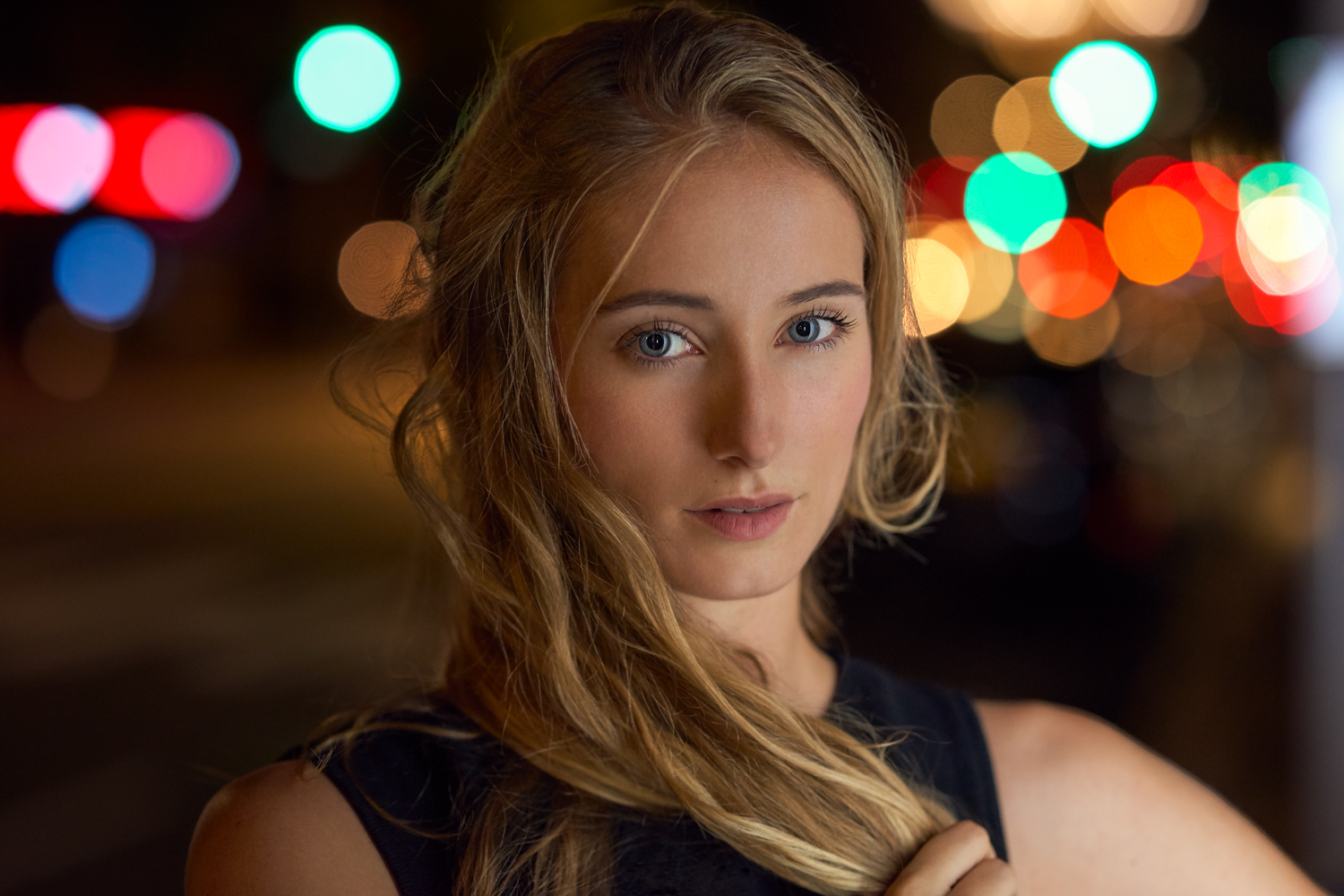 Portrait of a blonde woman with soft waves in her hair, set against colorful night bokeh. She looks calmly into the camera, lit with warm, flattering highlights.