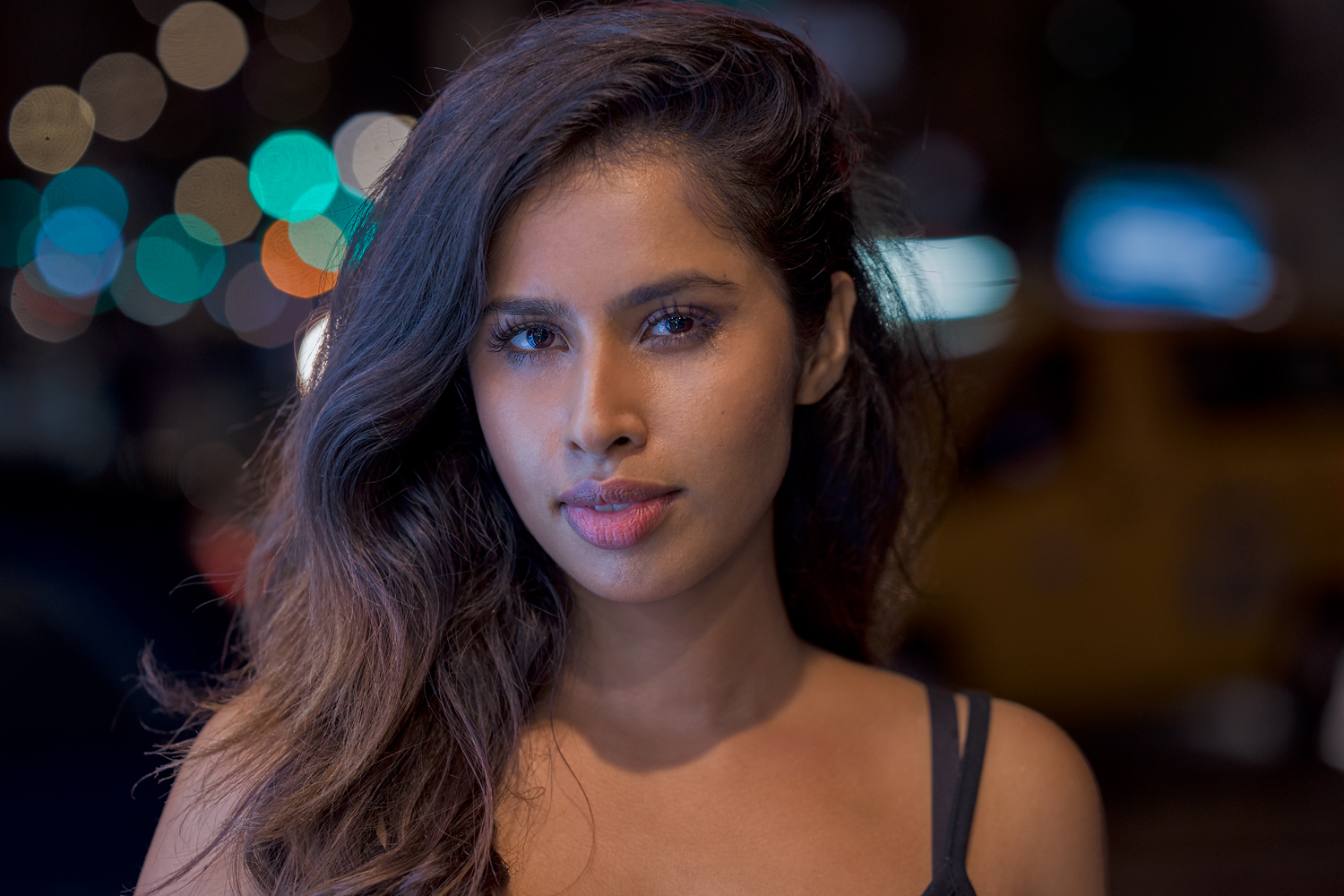 Portrait of a woman with long, wavy dark hair standing against a blurred city night backdrop. She gazes confidently into the camera, lit by soft, cinematic lighting.