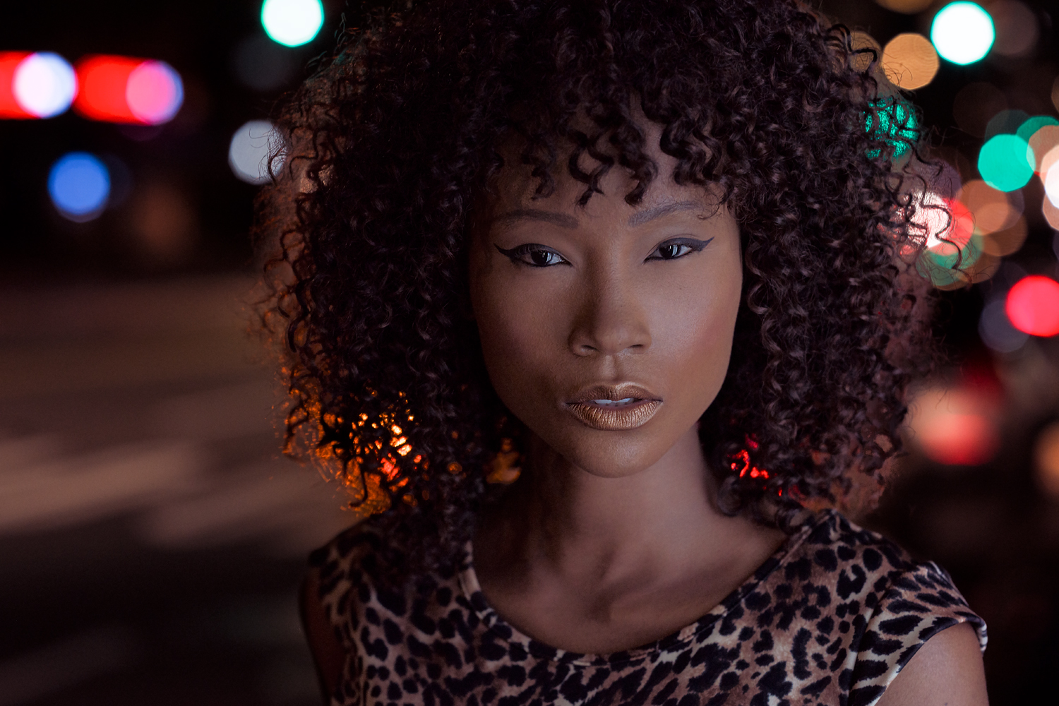 Portrait of a woman with voluminous curly hair wearing a leopard-print top, standing confidently against a colorful urban night backdrop with blurred city lights.