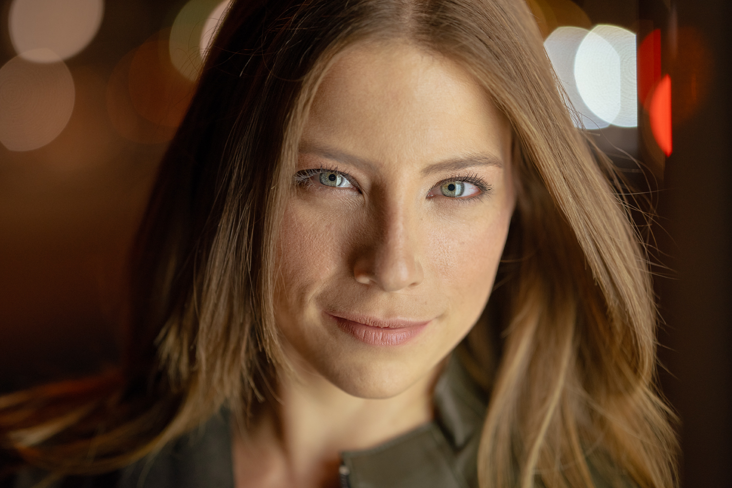 Portrait of a woman with light brown hair gazing directly at the camera, softly lit against a colorful, out-of-focus nighttime city background.