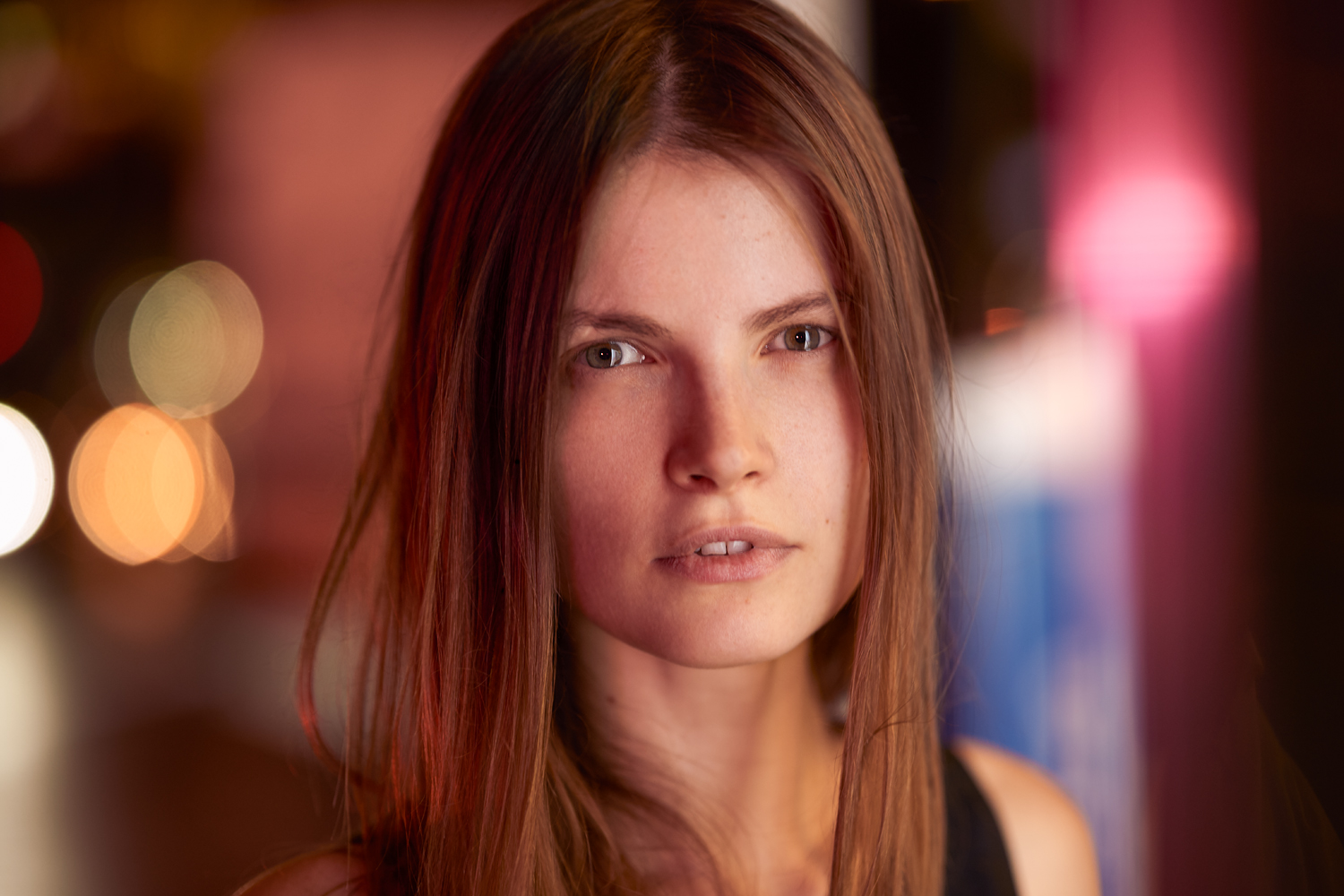 Portrait of a young woman with long brown hair, standing against a night city backdrop with colorful bokeh lights; soft red and amber lighting cast across her face.