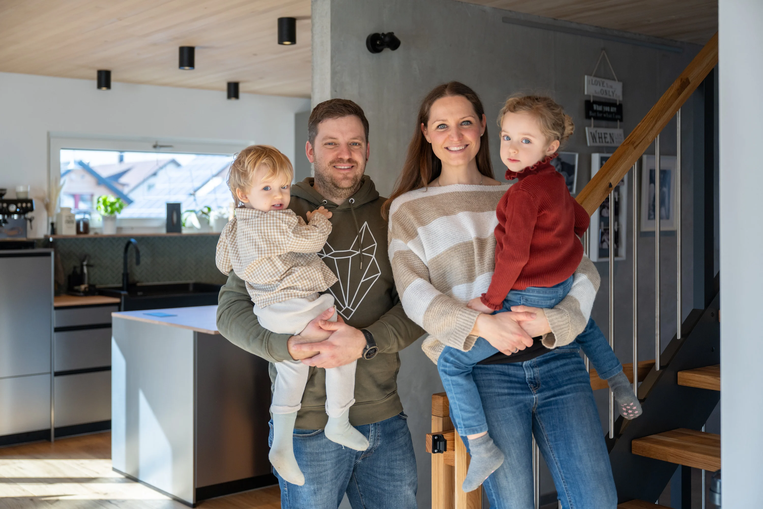 Familie mit Kindern auf den Armen in ihrem HAUSER-Haus vor Betonwand und Holztreppe.