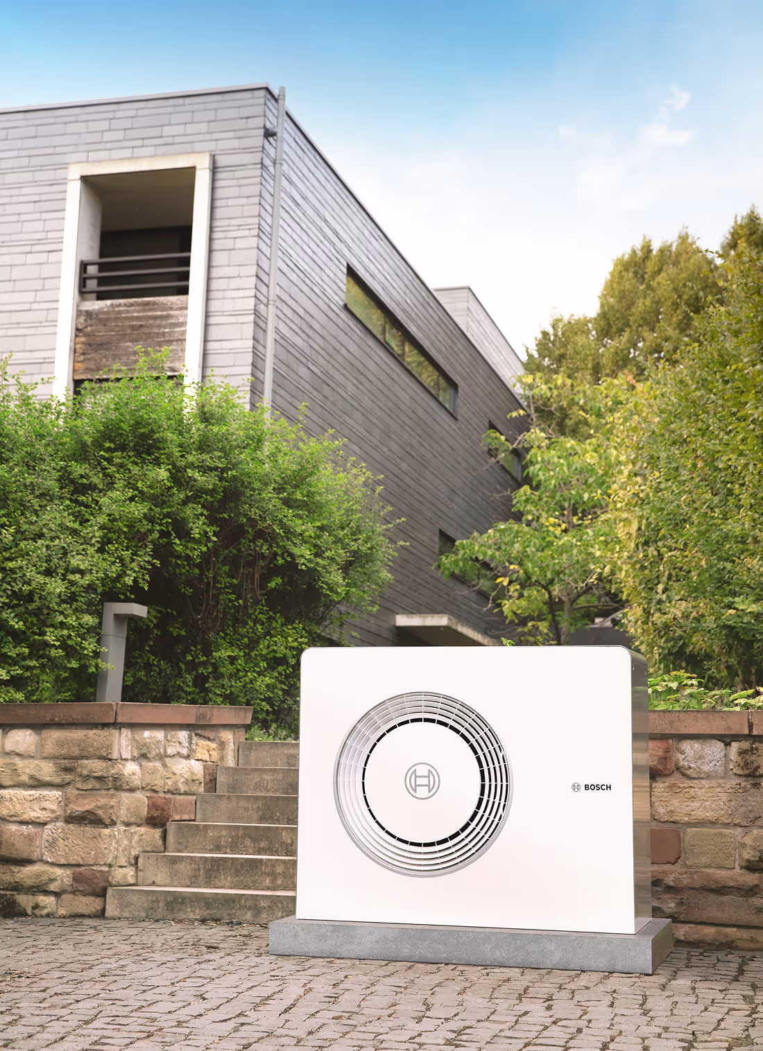 Bosch Wärmepumpe vor einer Mauer. Im Hintergrund Neubau Haus