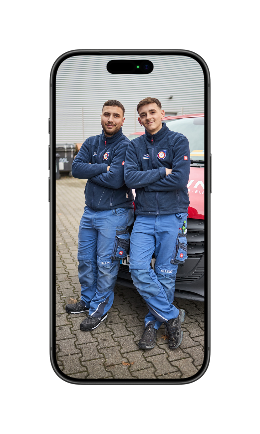 Zwei lächelnde Mitarbeiter stehen vor rotem Hintergrund; der Mann hält eine Rohrzange in der Hand.