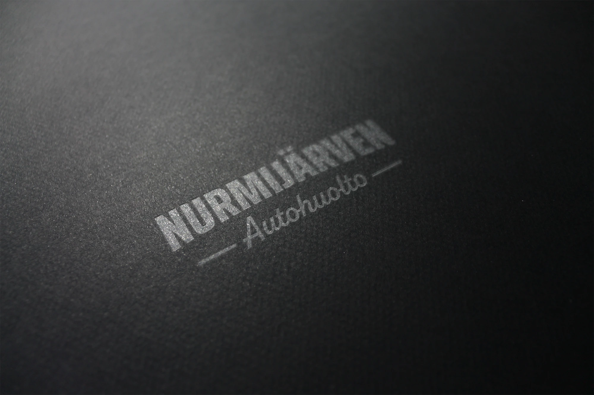 Nurmijärven autohuollon logo printattuna tumalle paperille hopeisena.