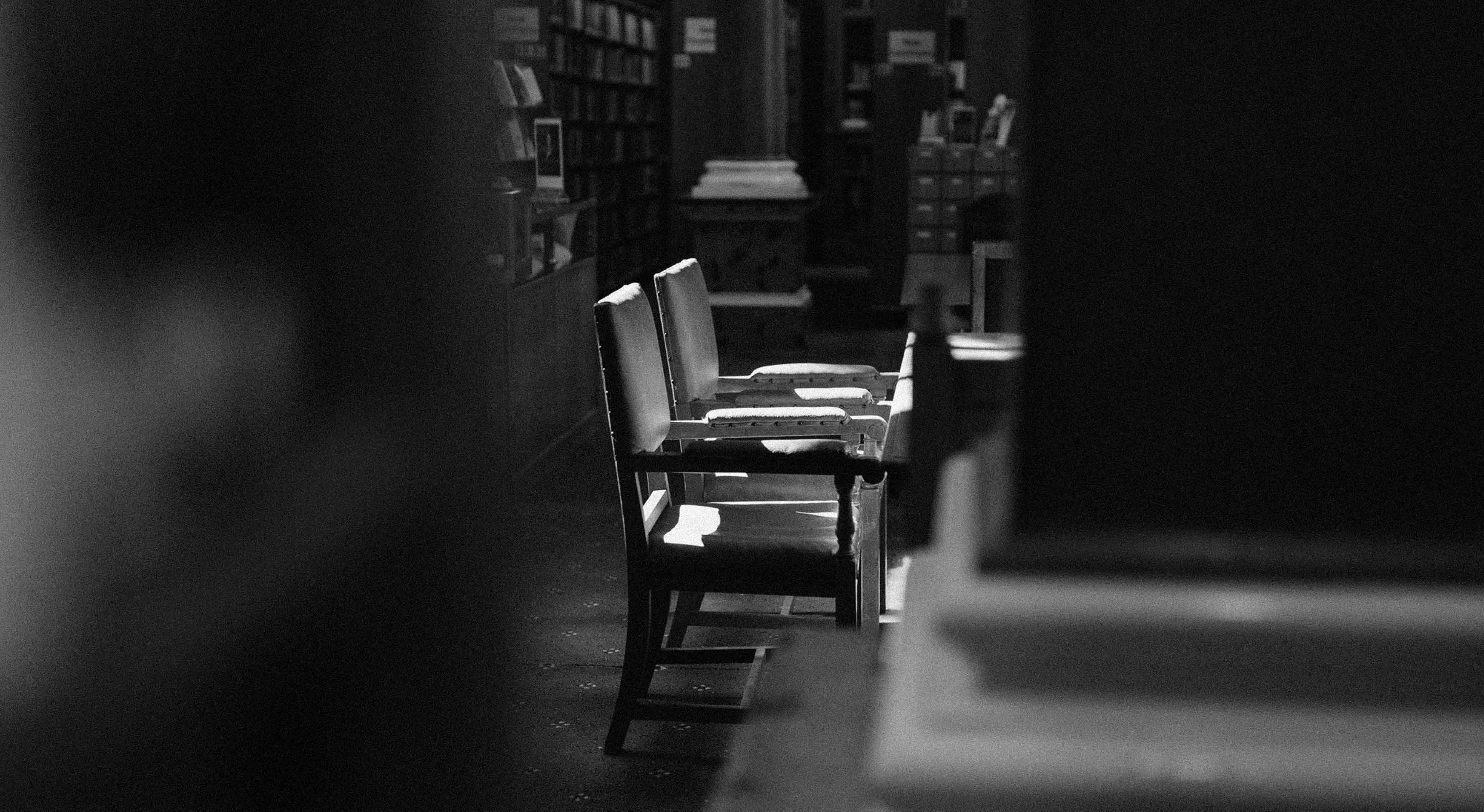 Three empty chairs at a library table with soft light highlighting their edges in a black and white photo.