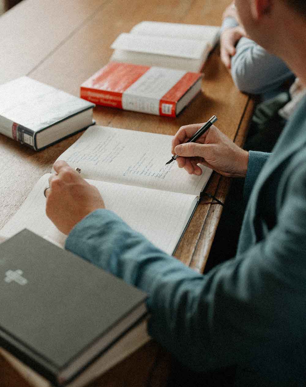 Person writing notes in an open notebook on a wooden table surrounded by several thick books.